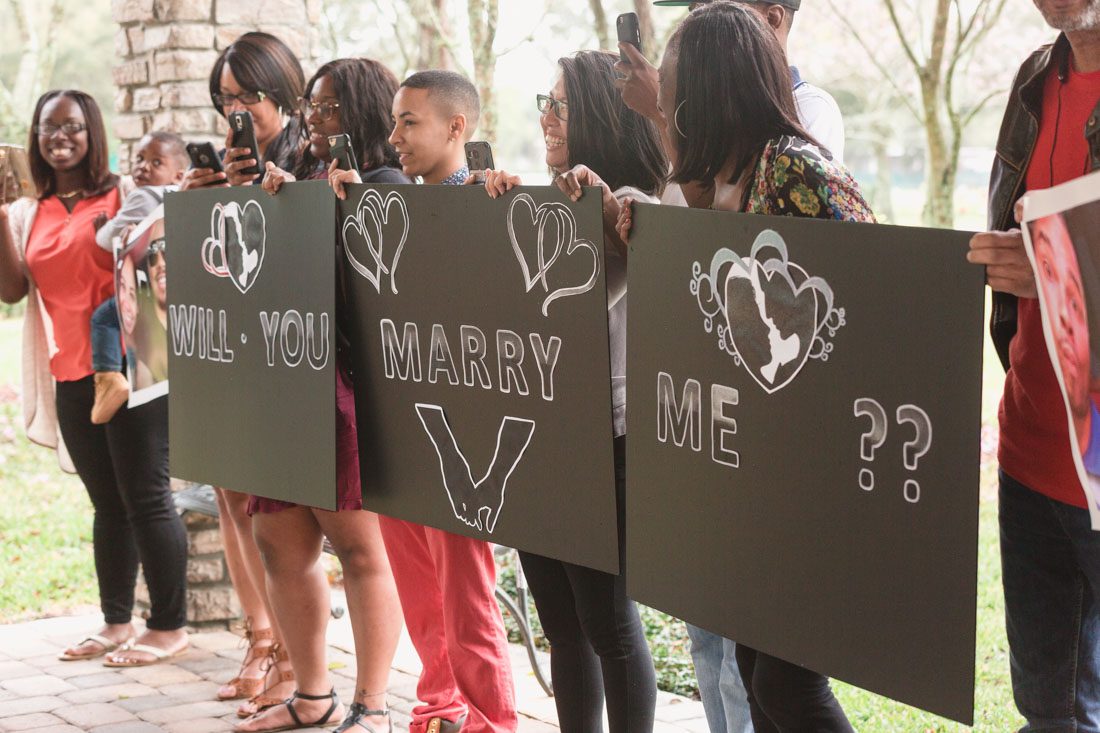 Family and friends hold signs and read will you marry me in a surprise proposal