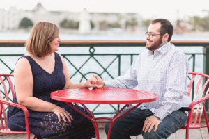 Newly engaged couple sit at a red bistro table overlooking the lake during their Disney Boardwalk engagement photography shoot