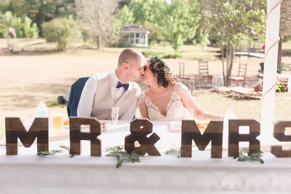 Newlyweds share a kiss at their sweetheart table featuring wooden Mr and Mrs signs during their outdoor reception