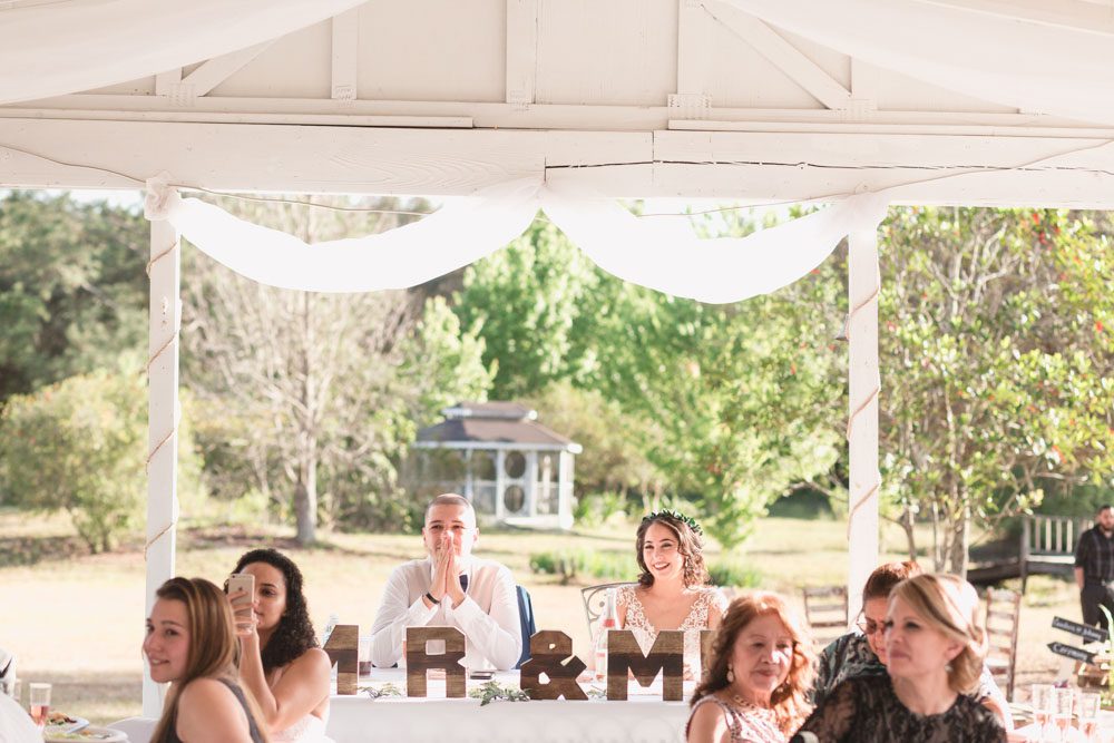 Bride and groom receive speeches from behind their sweetheart table during a romantic outdoor wedding reception in Orlando captured by top Central Florida wedding photographer